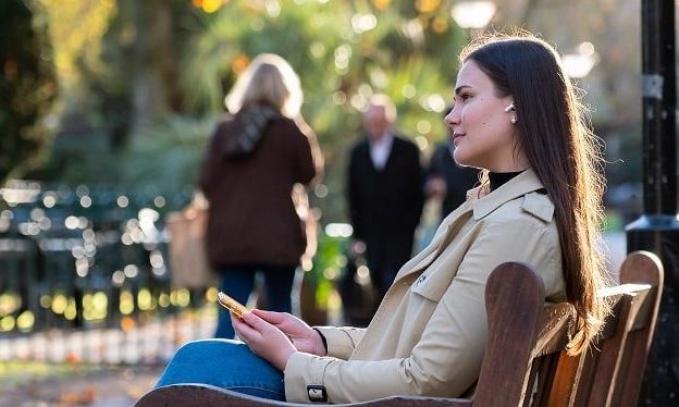 A young professional woman with headphones on is sat outside on a bench.