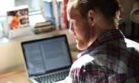 A photo of a man sitting by a laptop by the window